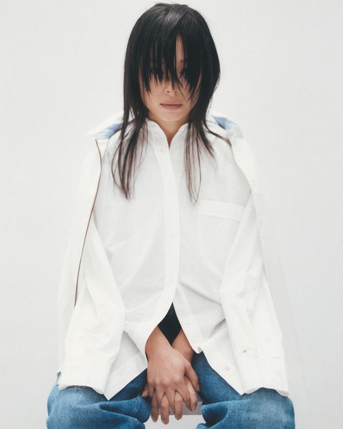 Asian woman with dark hair and blue jeans wearing a white button-down shirt poses in front of a white background.