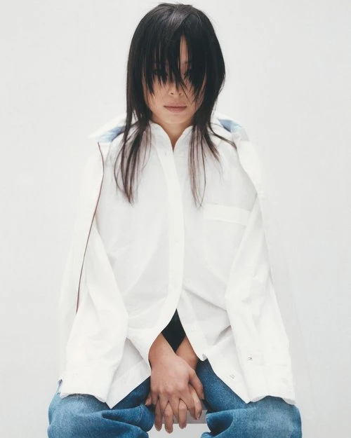 Asian woman with dark hair and blue jeans wearing a white button-down shirt poses in front of a white background.