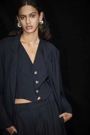 Fashionable woman in a dark vest and jacket poses against a black background, showcasing her modern and elegant style.