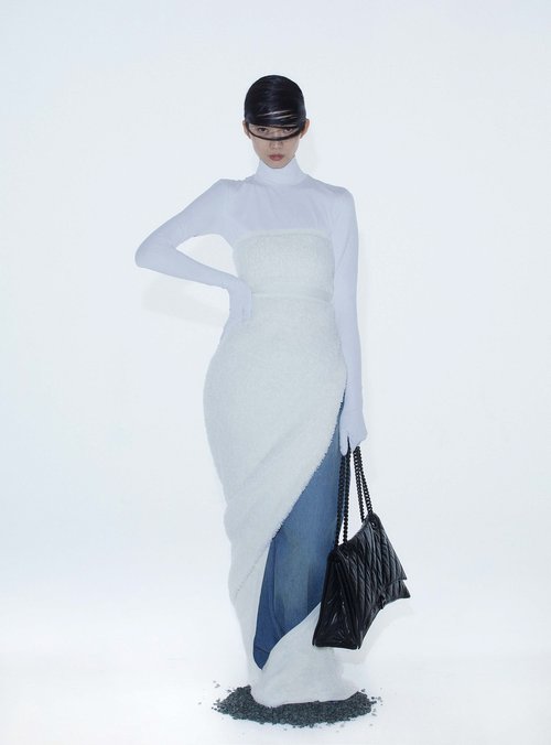 A slender woman in a futuristic white dress poses against a stark white backdrop, holding a black quilted purse. Fashion editorial.