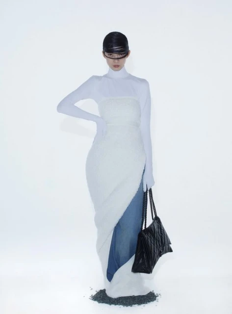 A slender woman in a futuristic white dress poses against a stark white backdrop, holding a black quilted purse. Fashion editorial.