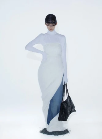 A slender woman in a futuristic white dress poses against a stark white backdrop, holding a black quilted purse. Fashion editorial.