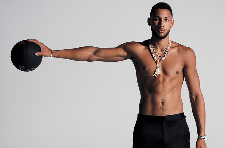 Shirtless male athlete, holding a black basketball with outstretched arm, showcasing his physique against a plain white backdrop.