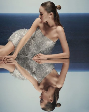 Model in strapless fringe dress reclining on a mirrored surface against an ocean horizon