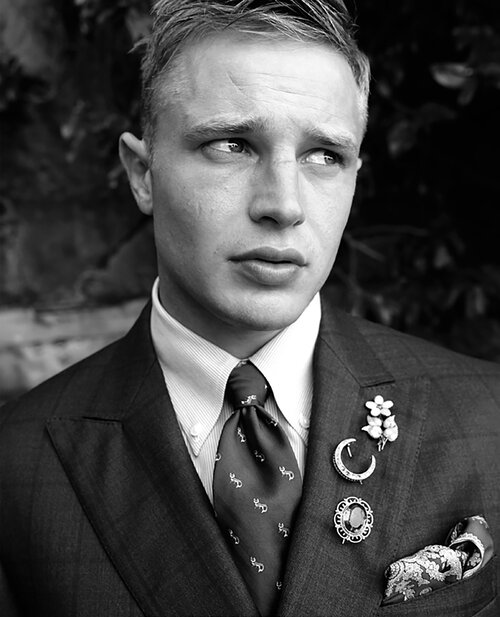 Monochrome close-up of a blond adult male in a dark suit, collared shirt, and patterned tie, accessorized with multiple brooches and a pocket square.