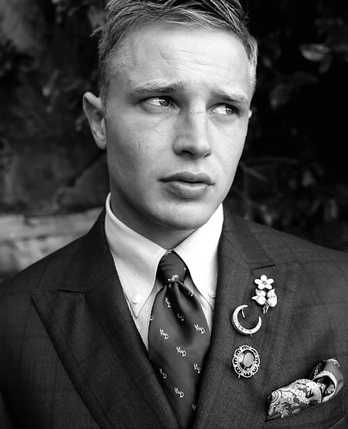 Monochrome close-up of a blond adult male in a dark suit, collared shirt, and patterned tie, accessorized with multiple brooches and a pocket square.