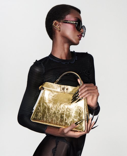 Fashionable Black model presents golden Fendi handbag with black manicure in studio shot with Fendi sunglasses and black long-sleeve dress.