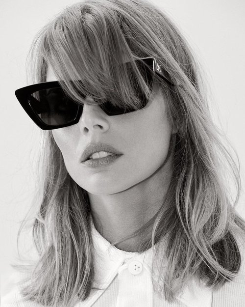Black and white portrait of an adult woman with blonde hair wearing black sunglasses and a white button-down shirt in studio lighting.