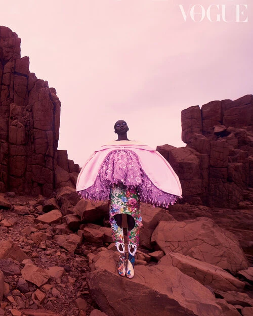 A dark-skinned model poses on a rocky landscape wearing a designer dress with pink accents for a Vogue fashion editorial.