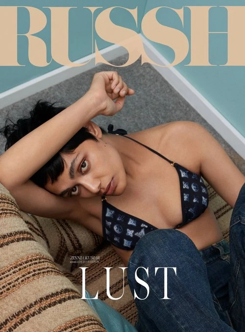 Model with dark hair wearing a patterned bikini top and blue jeans, posing indoors near a striped sofa by Pete Lennon.