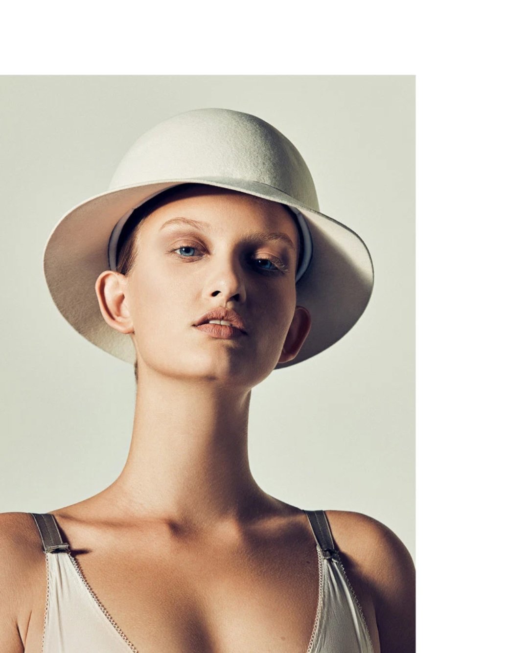 Close-up of a woman wearing a light colored hat, looking at the camera with a neutral expression, in a studio setting with soft lighting.