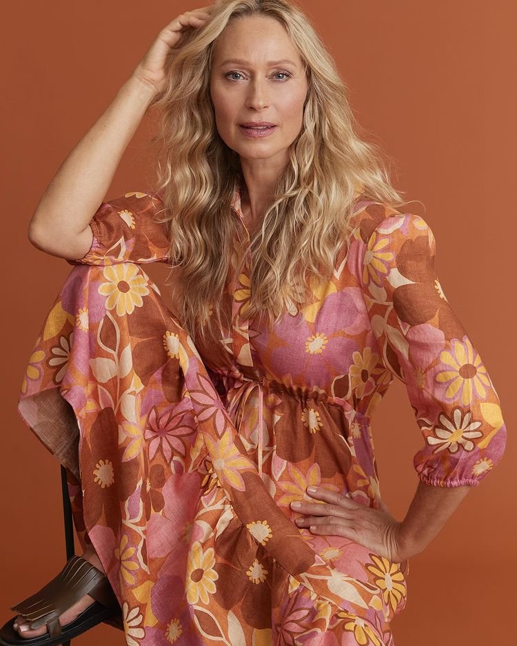 Blonde woman in a 70s-style floral dress posing indoors with an orange background. Portrait shot, eye-level angle, fashion photography.