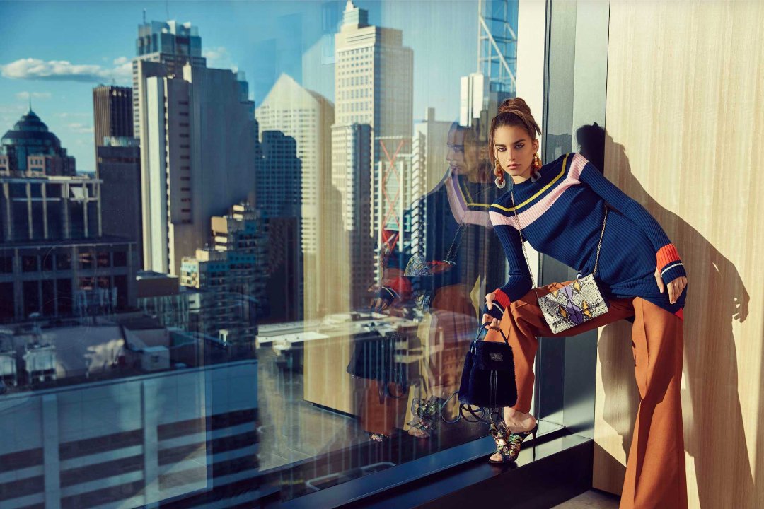 Fashionable woman in a blue dress and tan pants poses near a window with a city view, accessorized with handbags and statement earrings.