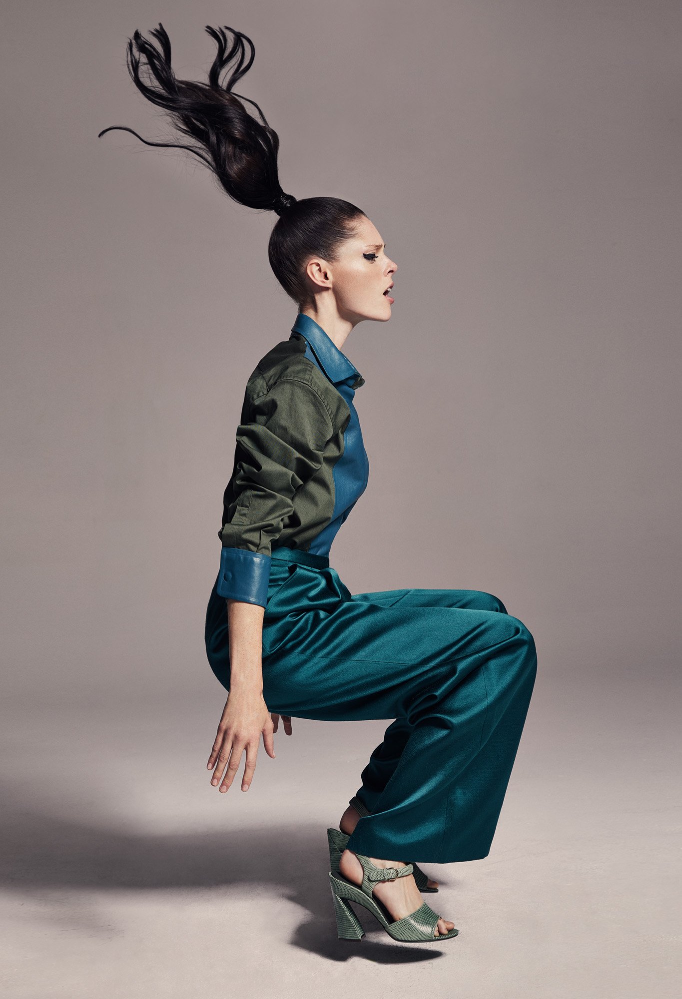 A woman with a high ponytail and green outfit jumps in a studio setting, highlighted by vibrant lighting and a grey background.