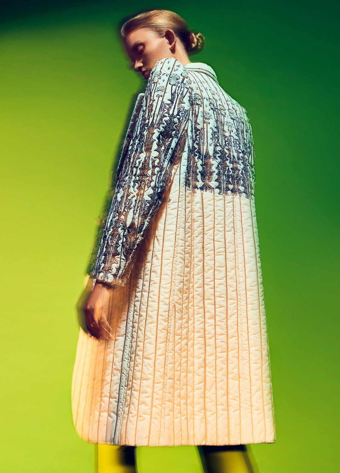 Woman in a long, detailed coat with light and dark patterned fabric on a green background, looking downwards, fashion forward, editorial.