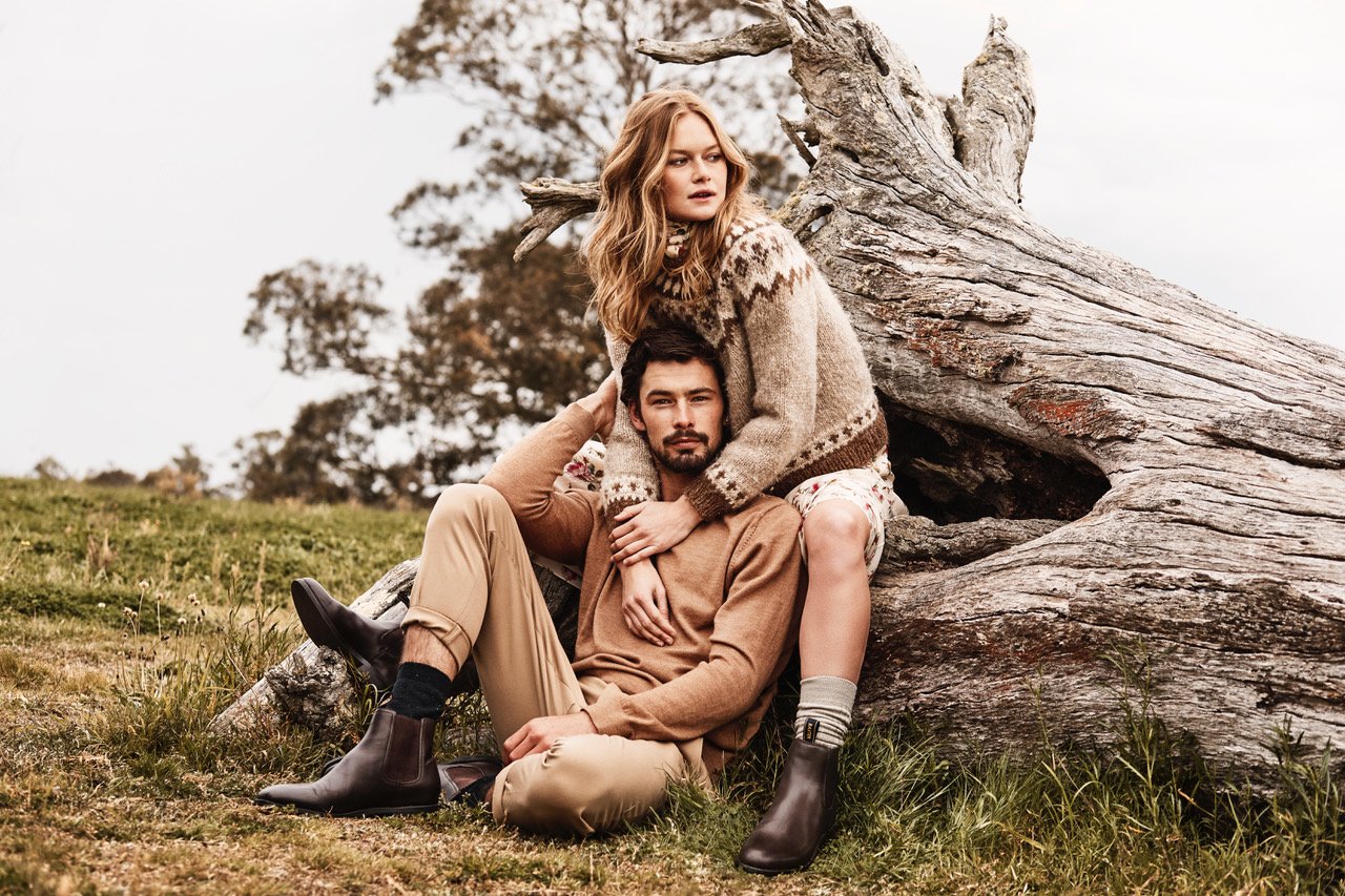 A stylish couple poses outdoors, the woman seated on a weathered log with her arms around the man, showcasing a fashion-forward fall look.