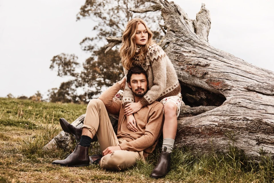 A stylish couple poses outdoors, the woman seated on a weathered log with her arms around the man, showcasing a fashion-forward fall look.