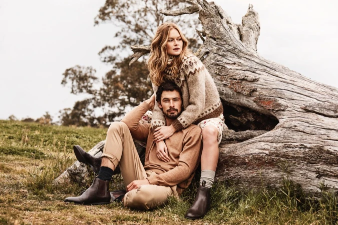 A stylish couple poses outdoors, the woman seated on a weathered log with her arms around the man, showcasing a fashion-forward fall look.