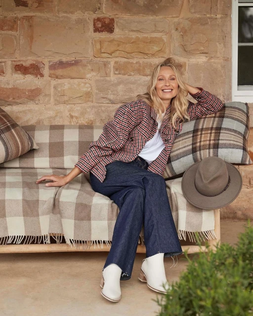Blonde woman in jeans and plaid jacket sits on a bench with pillows and a hat, smiling against a brick wall background.