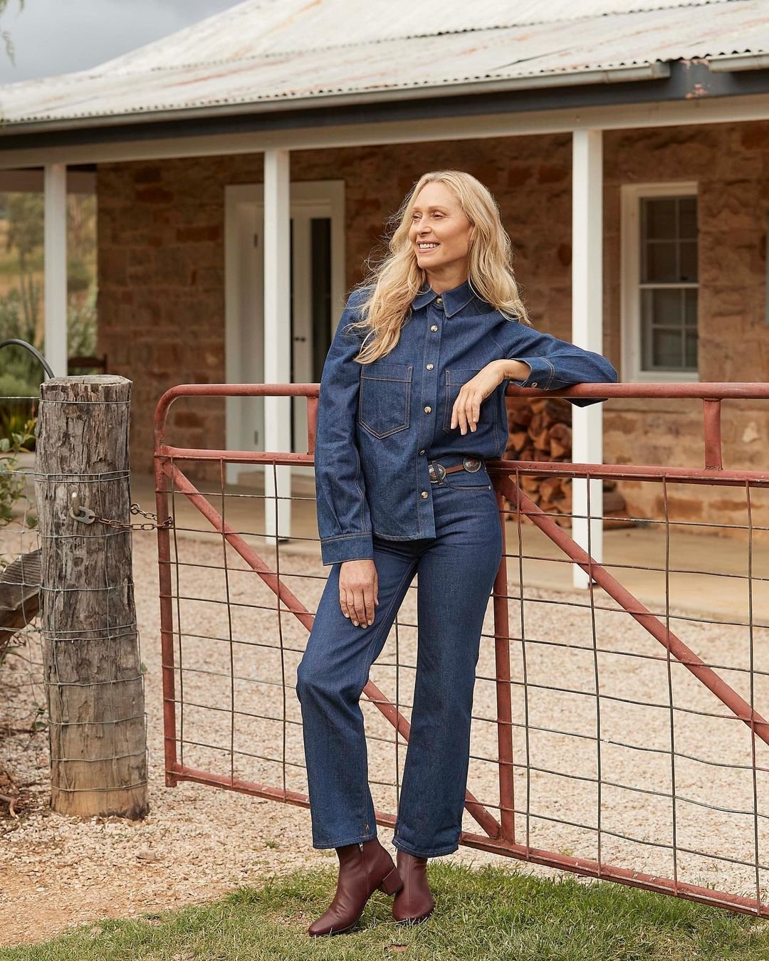 A blonde woman in a denim outfit leans against a metal gate in front of a stone house with green grass in the foreground.