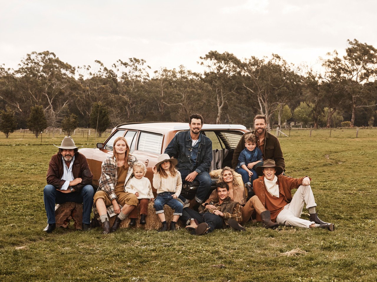 Group portrait of a large family gathered outside a brown station wagon on a grassy field, trees visible in the background, warm lighting.