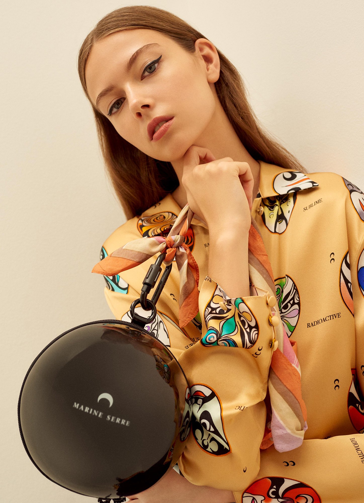 Young woman with long brown hair wearing a patterned top, holding a black spherical purse in an indoor setting. Fashion aesthetic.