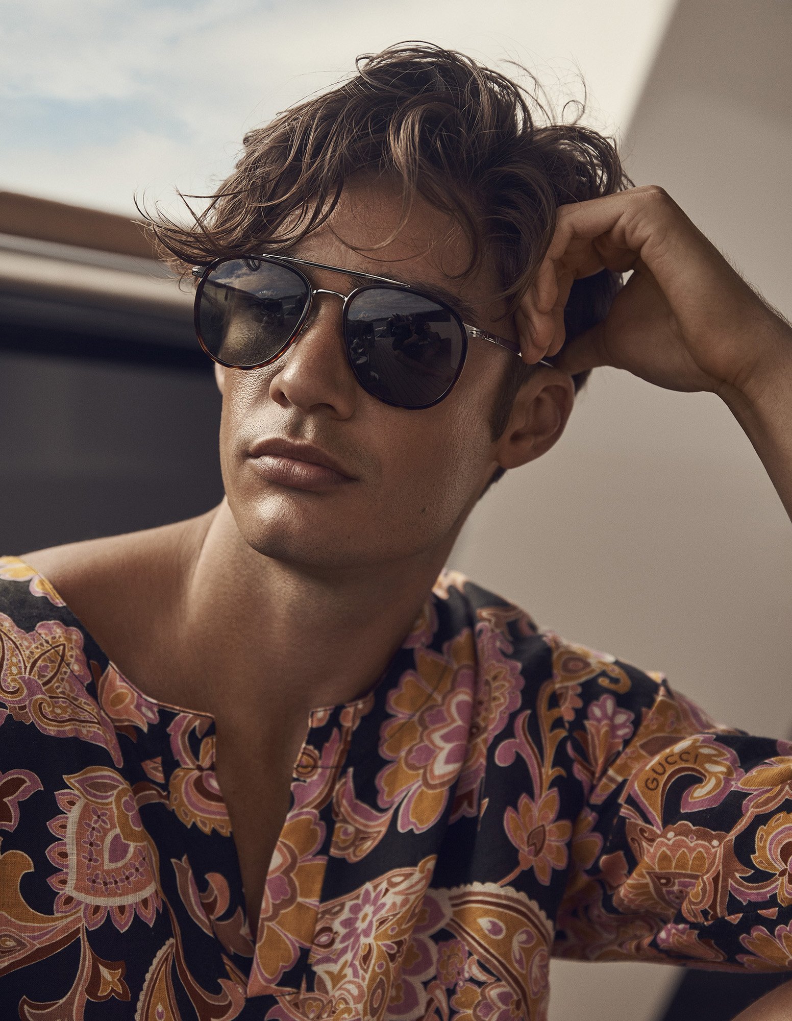 Close-up shot of a handsome adult male wearing sunglasses and a patterned top, lounging indoors with a neutral background and soft lighting.