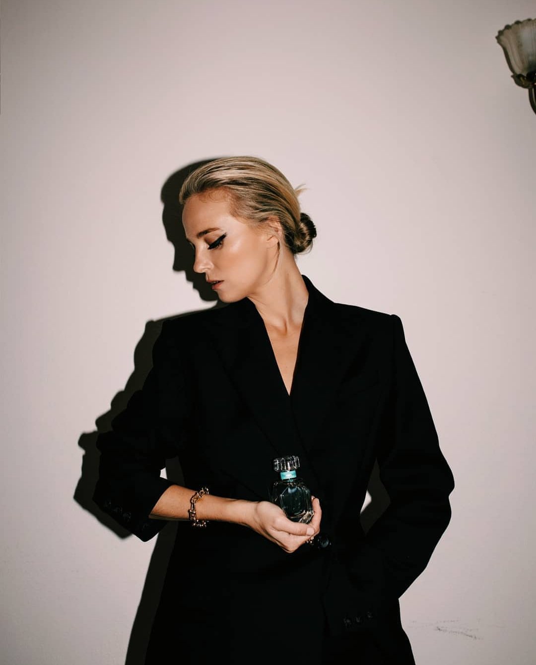 Blonde woman in a black blazer holding a perfume bottle against a neutral wall with strong shadow casting