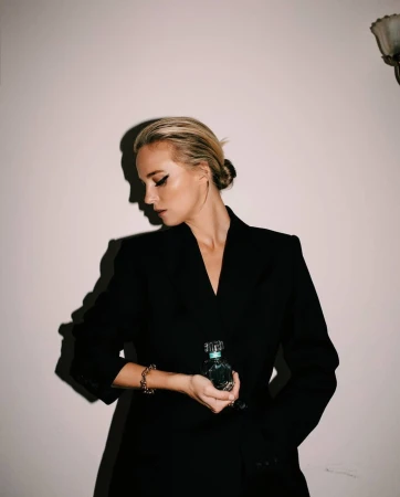 Blonde woman in a black blazer holding a perfume bottle against a neutral wall with strong shadow casting