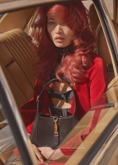 A red-haired woman in a car wearing a red jacket and holding a black handbag, illuminated by natural light and framed by the car's interior.