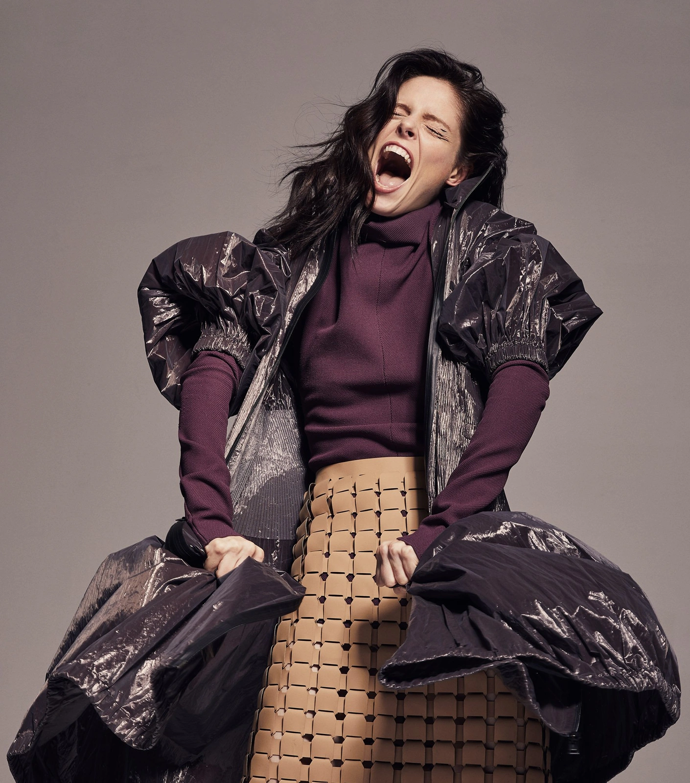 Model with dark hair and make-up by Elsa Morgan screaming with mouth open, wearing sculptural purple turtleneck and grid-pattern skirt.