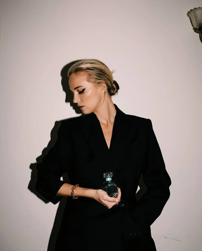 Blonde woman in a black blazer holding a perfume bottle against a neutral wall with strong shadow casting