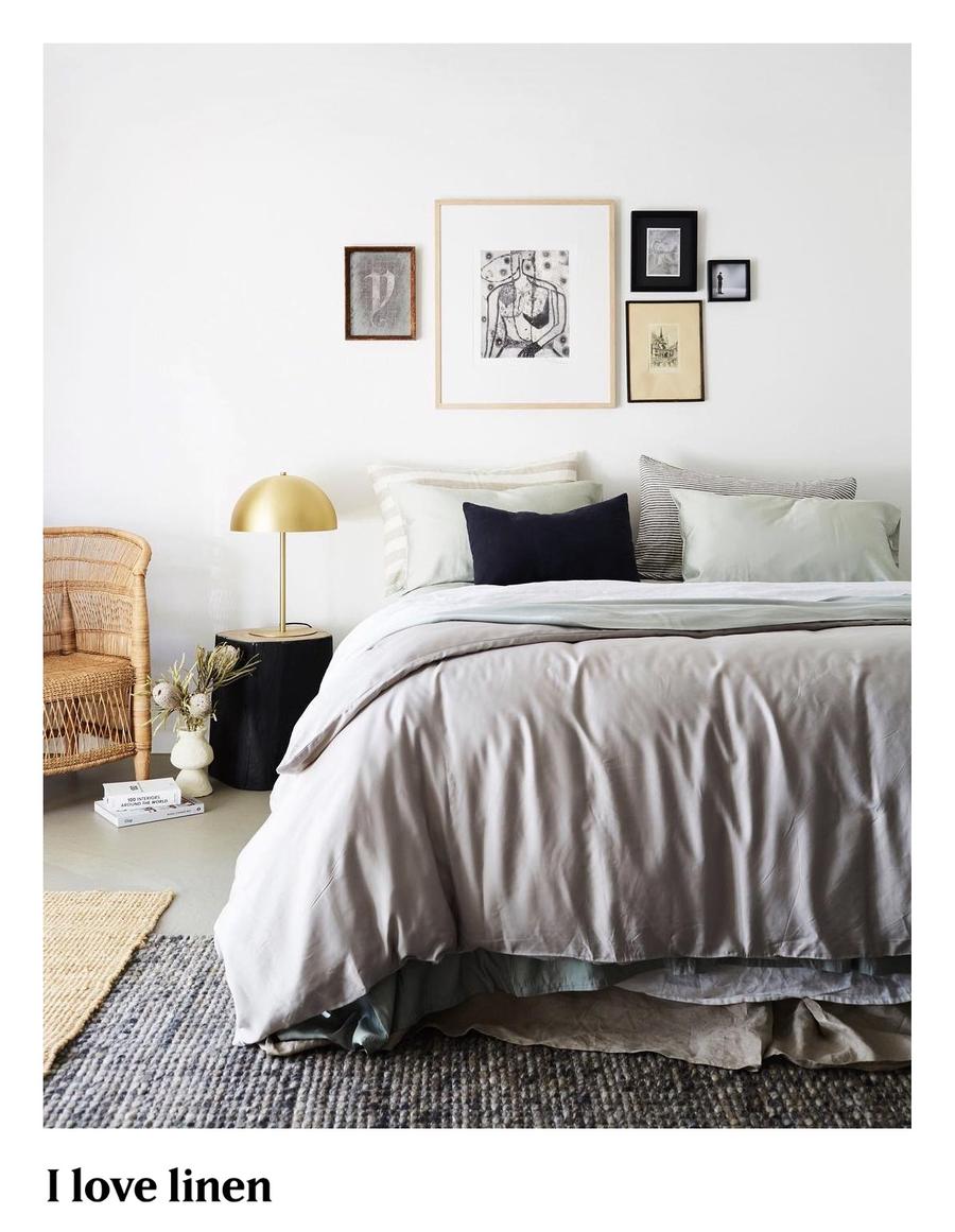 Bedroom interior featuring a bed with linen bedding, a wicker chair, a gold-colored lamp, and artwork on the wall in a minimal setting.