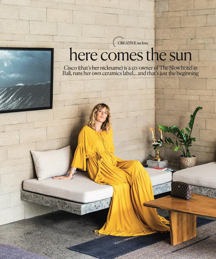 Woman in long yellow dress sits on a bench in a modern indoor setting with a framed art print and table. Magazine editorial layout.