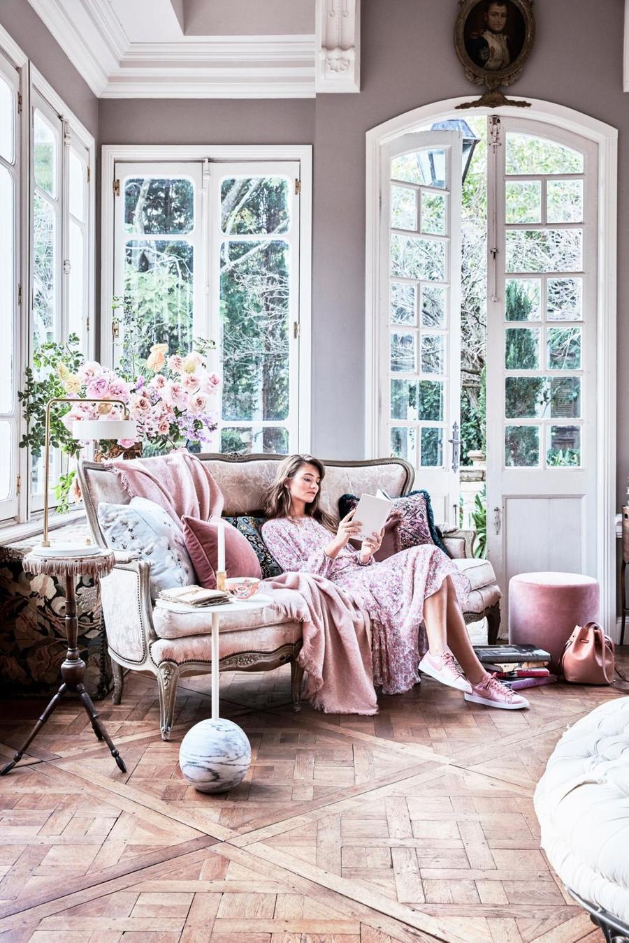 Woman lounges on an ornate couch in a sunlit room, reading. The indoor space features large windows, parquet floors, and pastel pink accents.