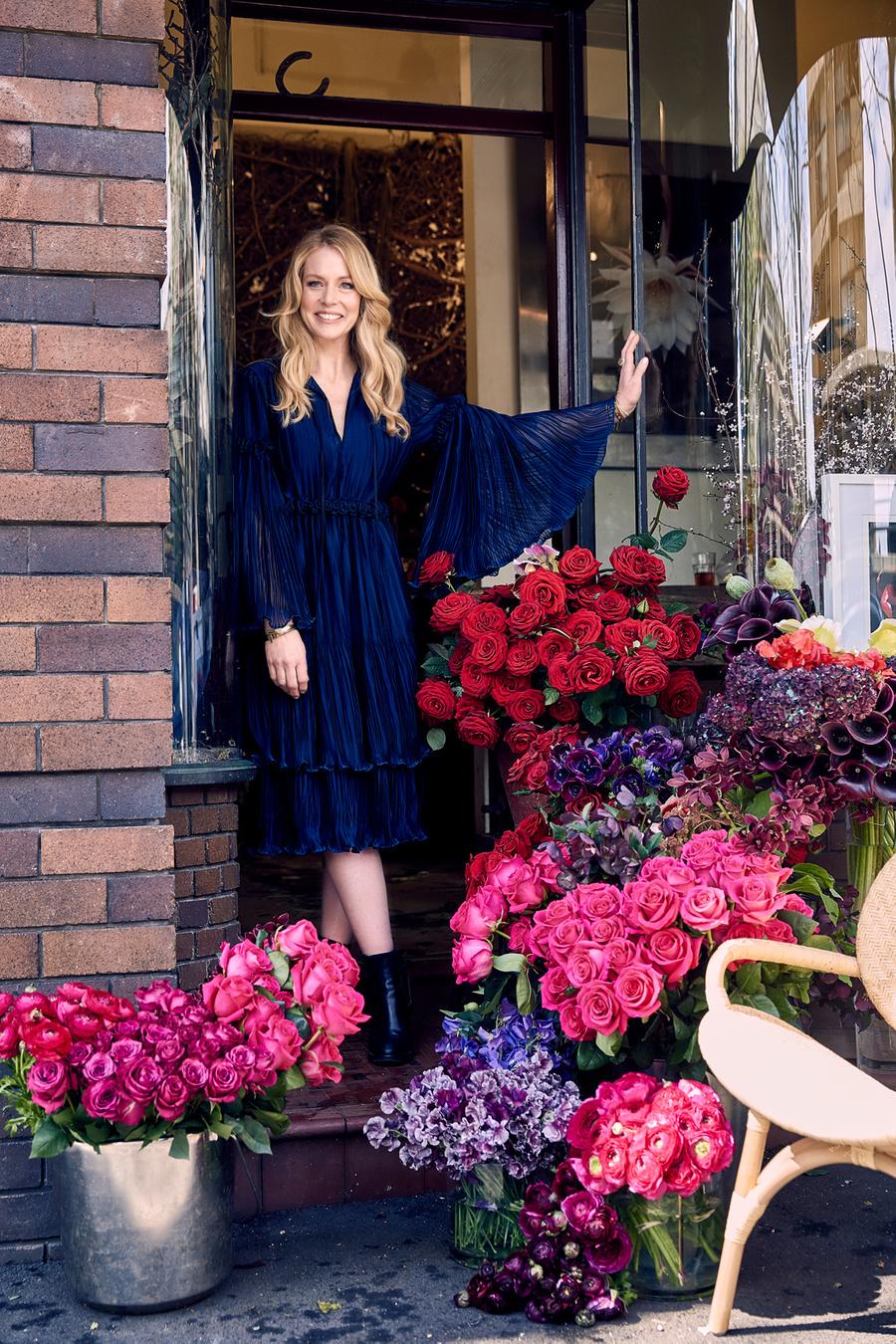 Blonde woman poses in a navy blue dress in front of a flower shop with red and pink roses in buckets and vases outside the entrance.