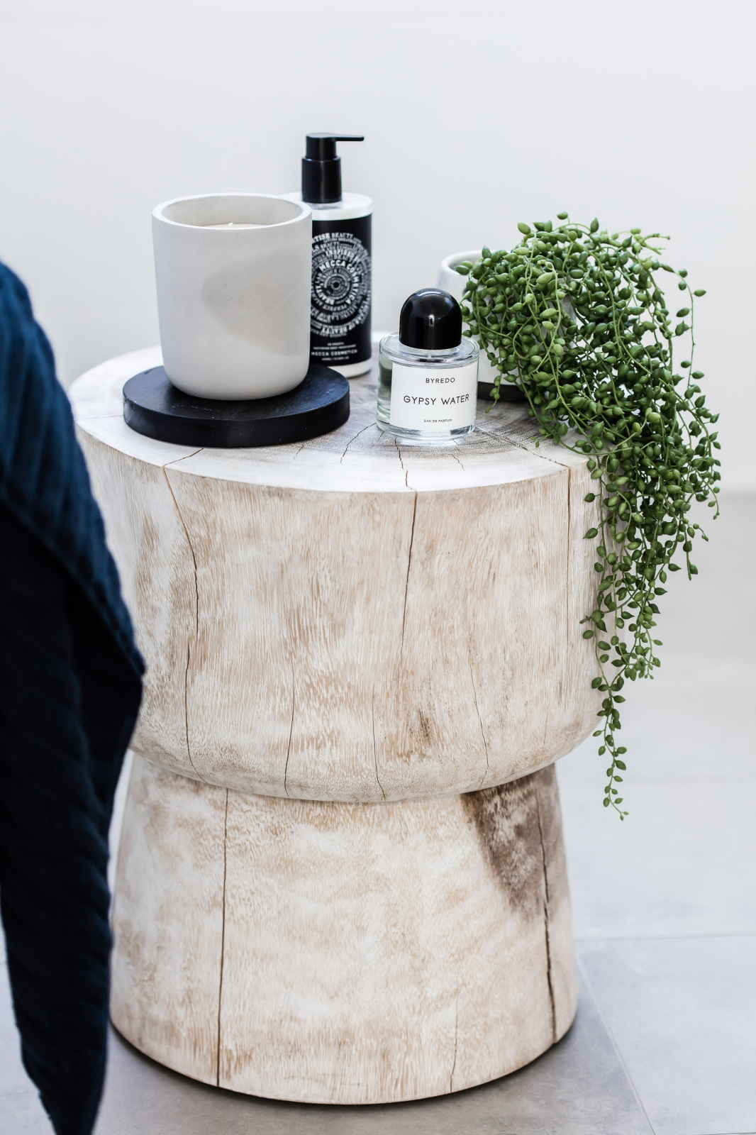 A wooden side table displaying a candle, soap dispenser, perfume bottle, and trailing plant in a bright indoor setting, captured with neutral tones.