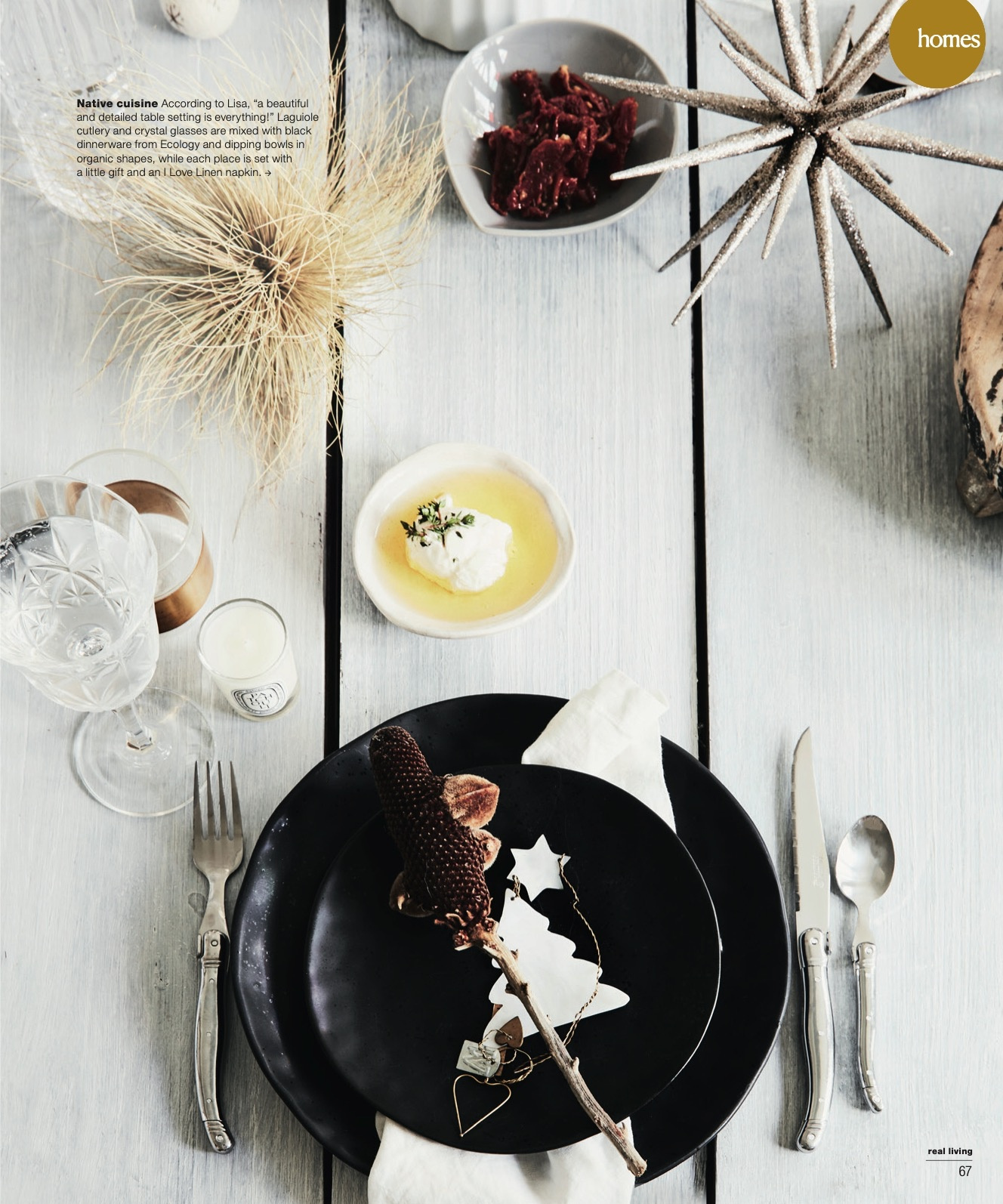 Overhead shot of a styled table setting with a black plate, silverware, and decorative items on a light-colored wooden surface.