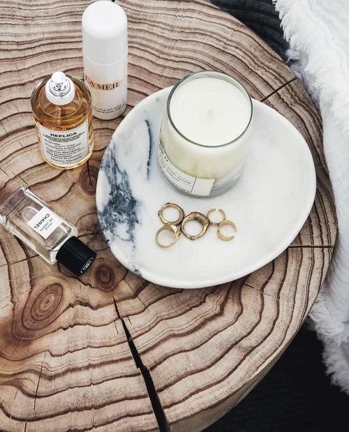 Still life of perfumes and a candle on a wood table. A white and marble tray holds rings and a white candle, perfumes are next to it.