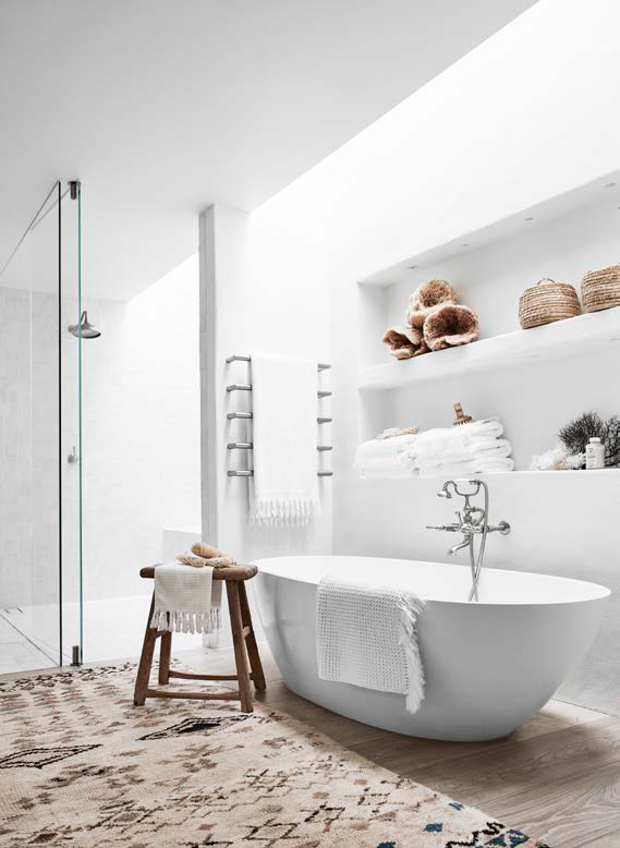 Bright bathroom with white, oval bathtub, patterned rug, and wooden stool with towels. A glass-enclosed shower stands nearby.