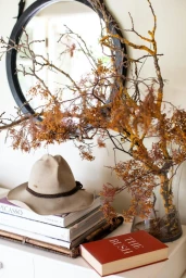 A cream-colored wide-brim hat resting on a stack of art books beside a glass vase with branches and a round black mirror on the wall.
