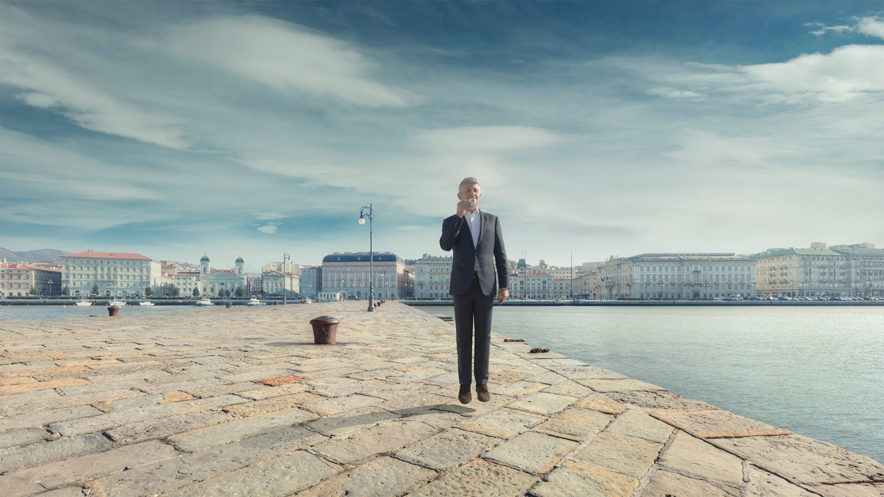 A man in a dark business suit is levitating on a stone pier with buildings and water in the background on a cloudy day.