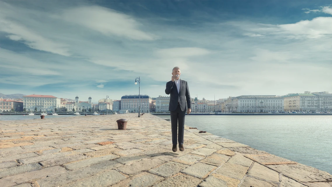 A man in a dark business suit is levitating on a stone pier with buildings and water in the background on a cloudy day.