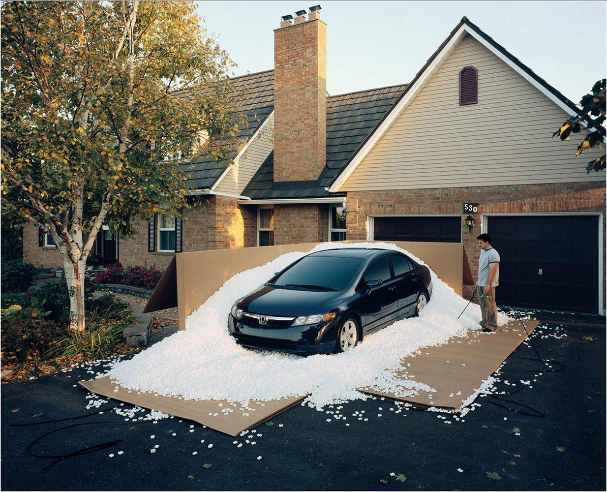 Car is half buried in styrofoam pellets on a driveway of a house with a man holding a hose in the background.