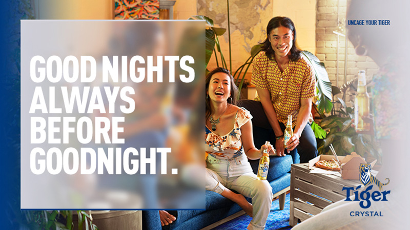 Two smiling adults, Asian ethnicity, seated inside with beer bottles, pizza, and plants, enjoying an evening with ambient indoor lighting.
