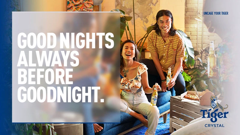 Two smiling adults, Asian ethnicity, seated inside with beer bottles, pizza, and plants, enjoying an evening with ambient indoor lighting.