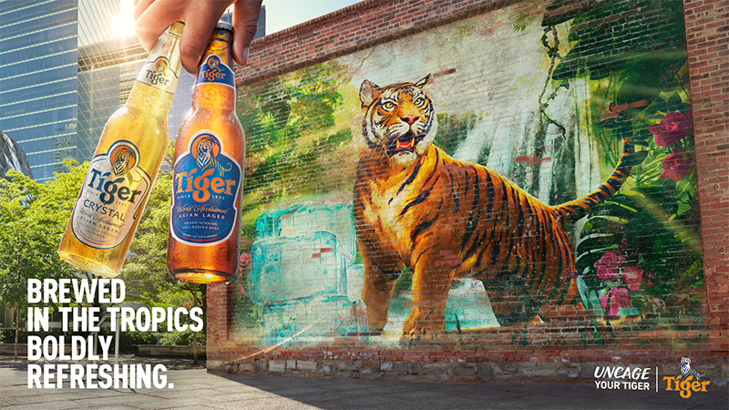 Two bottles of Tiger beer are held in front of a brick wall with a tiger mural, a city, and green trees. The text reads: Brewed in the tropics.