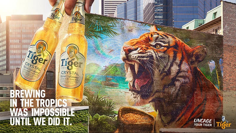 Two Tiger Crystal beer bottles held up in front of a mural of a tiger and a cityscape of tall buildings. Beer advertising.