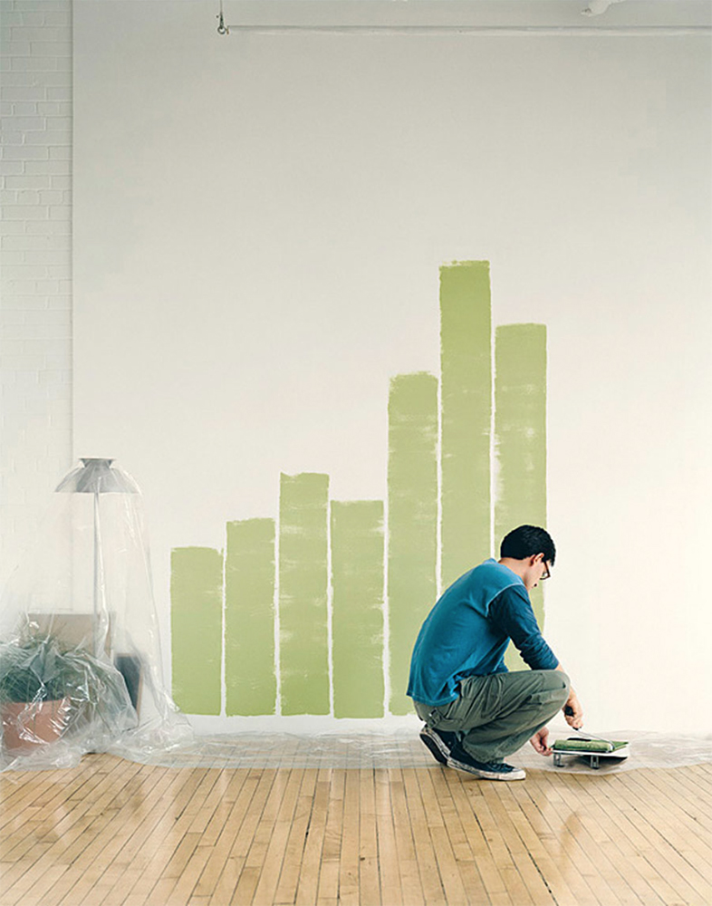 Man crouches painting a chart on an interior wall with light green paint. The room is a studio with wood floors.