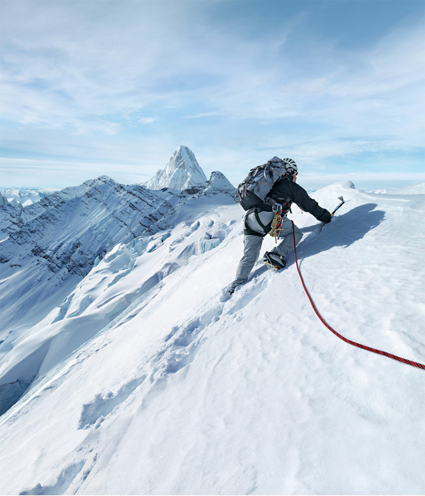 Adult male mountain climber with backpack ascends snow-covered mountain with ice axe, secured by red climbing rope under blue sky.